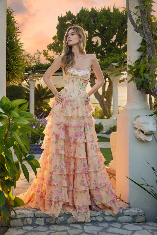 A woman stands outdoors at sunset before a garden pergola, wearing the Juliet JT2739S Long Prom Dress in Pastel Yellow Floral, featuring a strapless corset bodice and a flowing, ruffled chiffon A-line skirt with pink flowers.