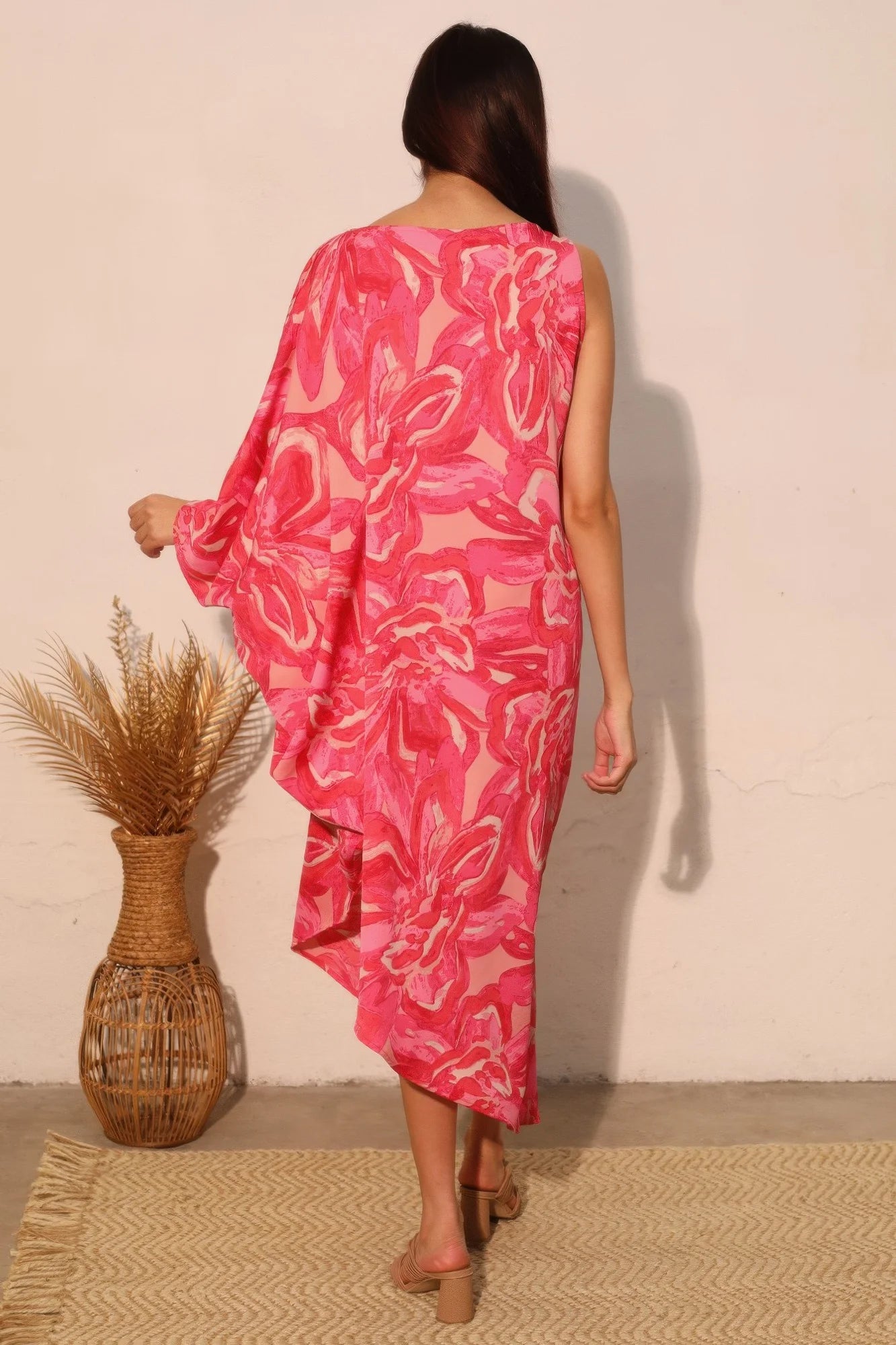 Woman wearing a pink floral dress standing against a beige wall with a woven basket and plant decor.