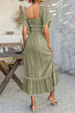 Woman wearing a green dress with ruffled sleeves and hem, standing in a room with a wooden table and decorative items.