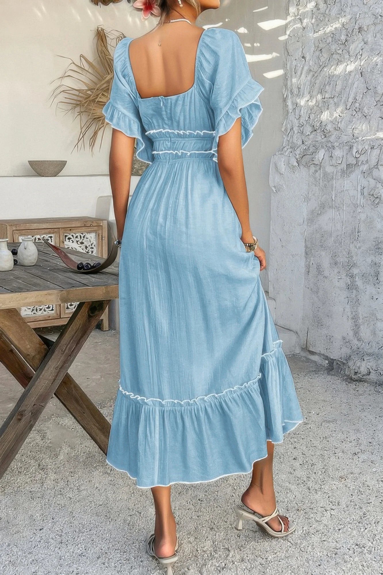 Woman wearing a light blue dress with ruffled sleeves and hem, standing in a room with a wooden table and decorative items.