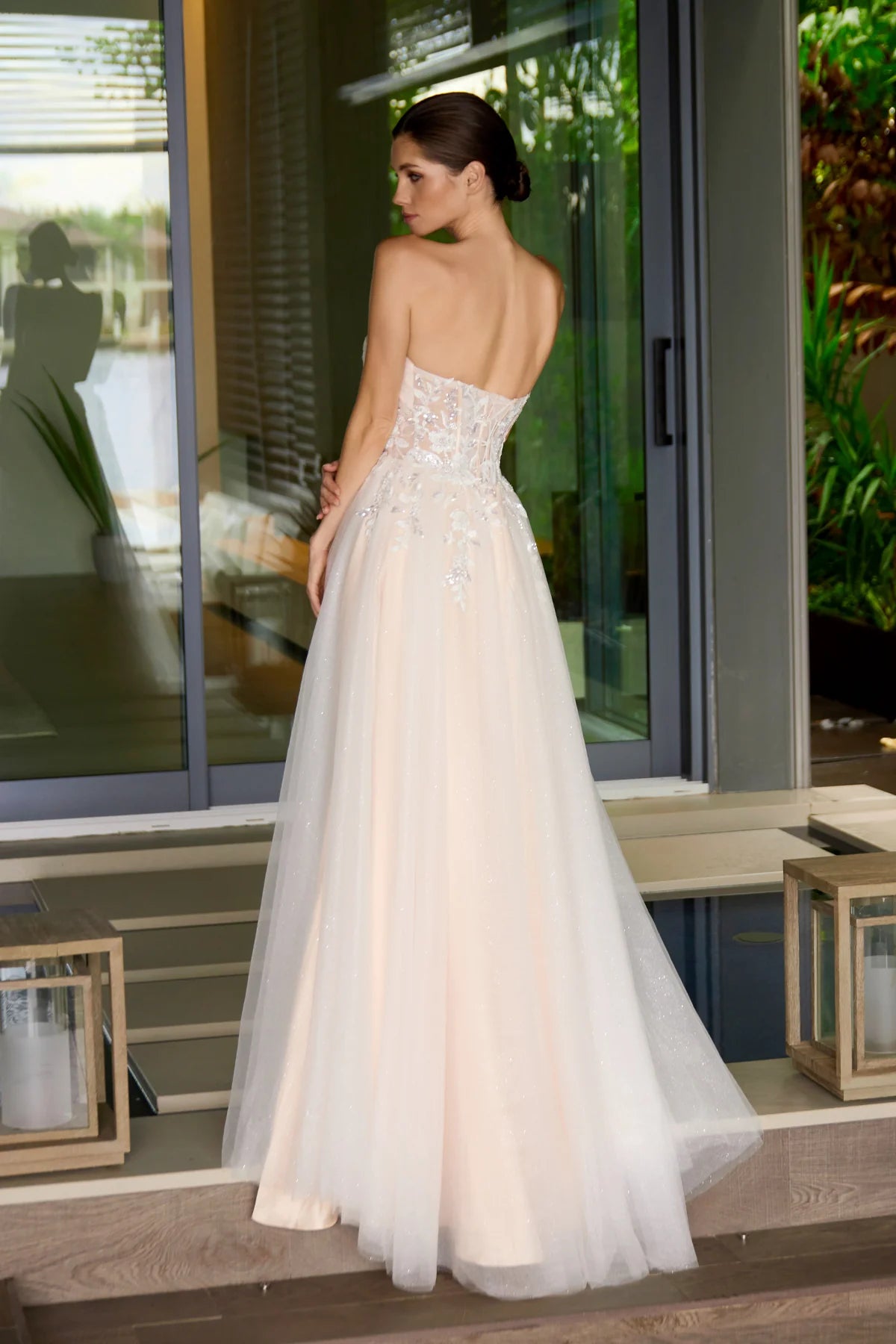 Woman in a nude strapless wedding gown standing in an outdoor setting with large windows and plants.