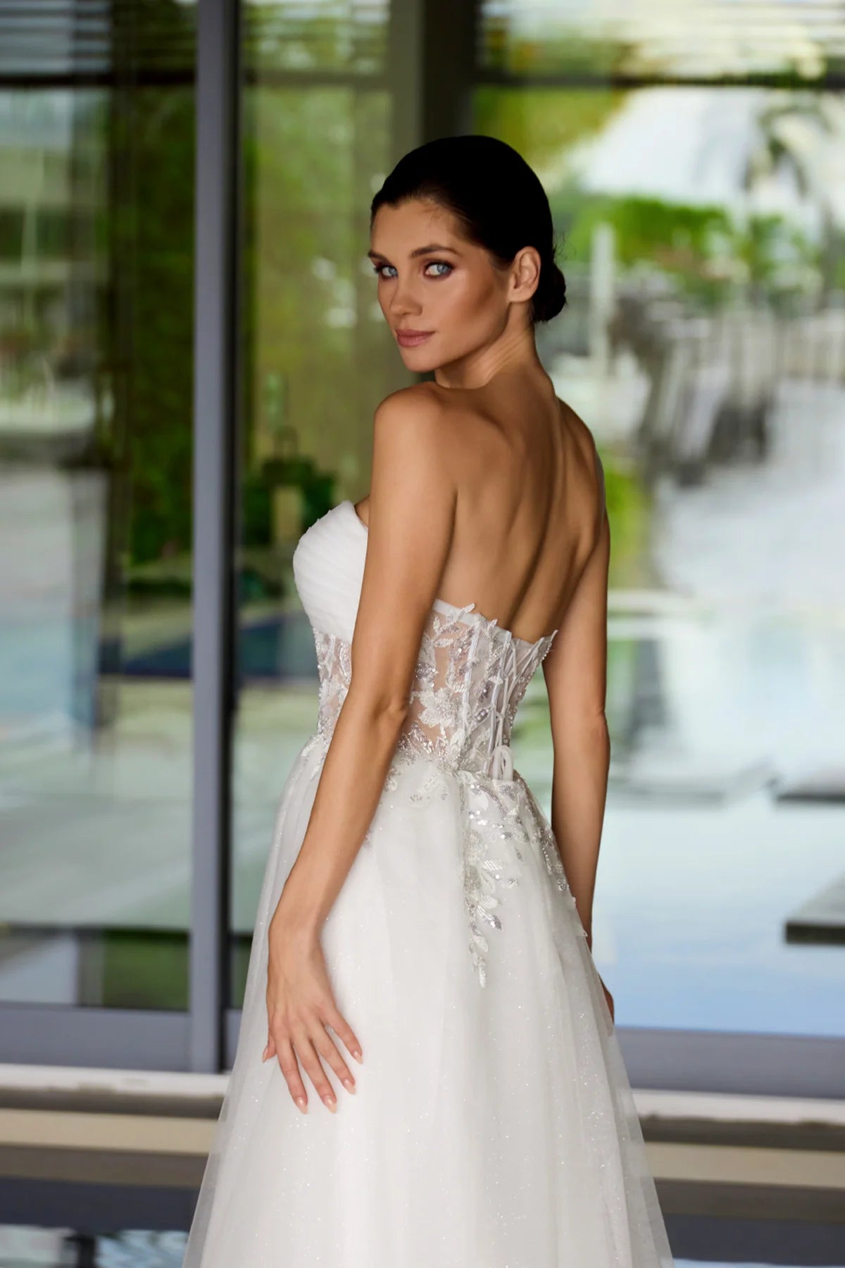 Woman in a white wedding dress with an open back, standing in front of a glass door.