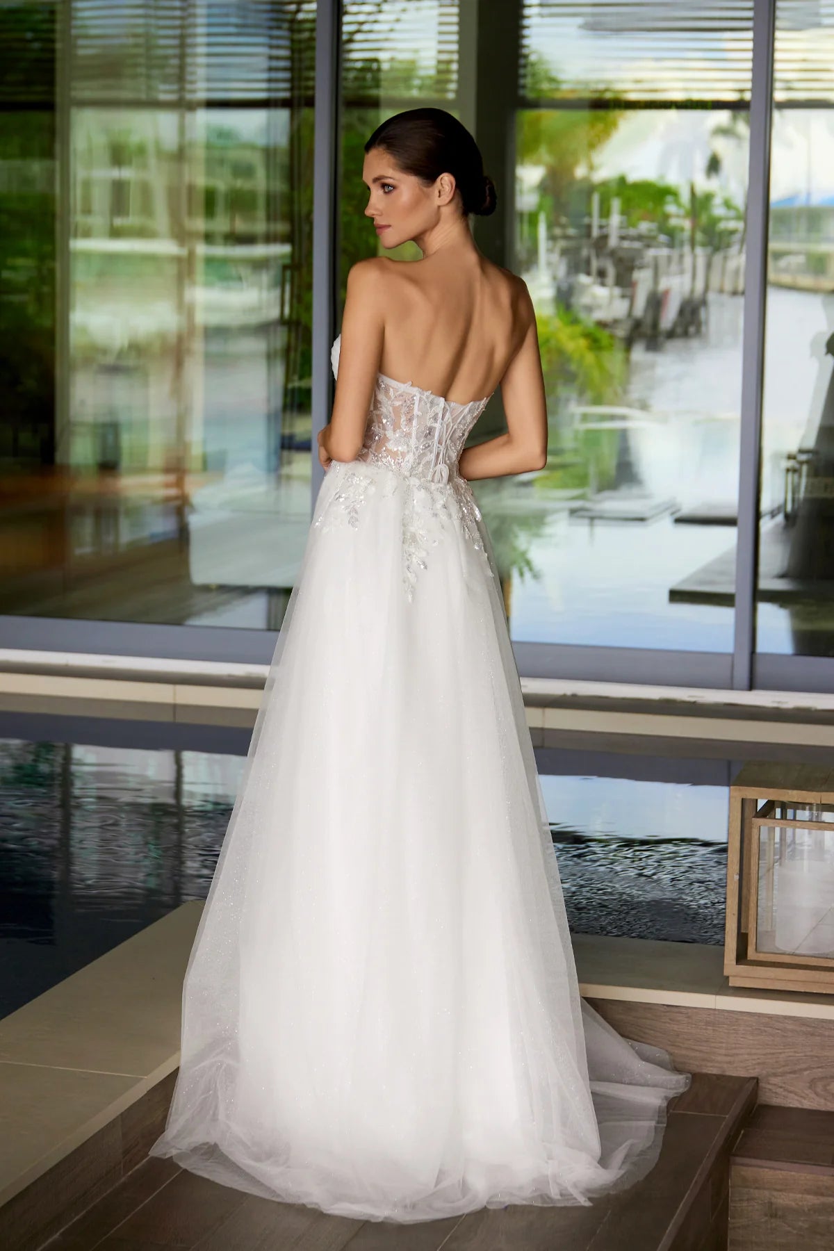 Woman in a white wedding dress standing in front of a large window with a view of greenery.
