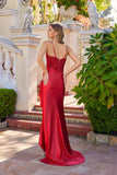 A woman wears the Juliet JT2697A long red prom dress with a thigh-high slit, spaghetti straps, and beaded corset bodice, standing outdoors amid greenery and stone owl statues.