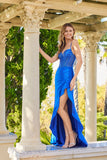 A woman in the Juliet JT2697A Long Prom Dress in royal blue with a satin skirt and beaded corset bodice stands outdoors by a stone column, smiling and posing with one hand resting on the column.