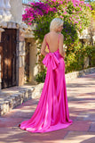 A woman with long blonde hair wears the Juliet JT2728H bright pink, floor-length corset prom dress with a large back bow, standing outdoors on a stone pathway surrounded by greenery and flowers.