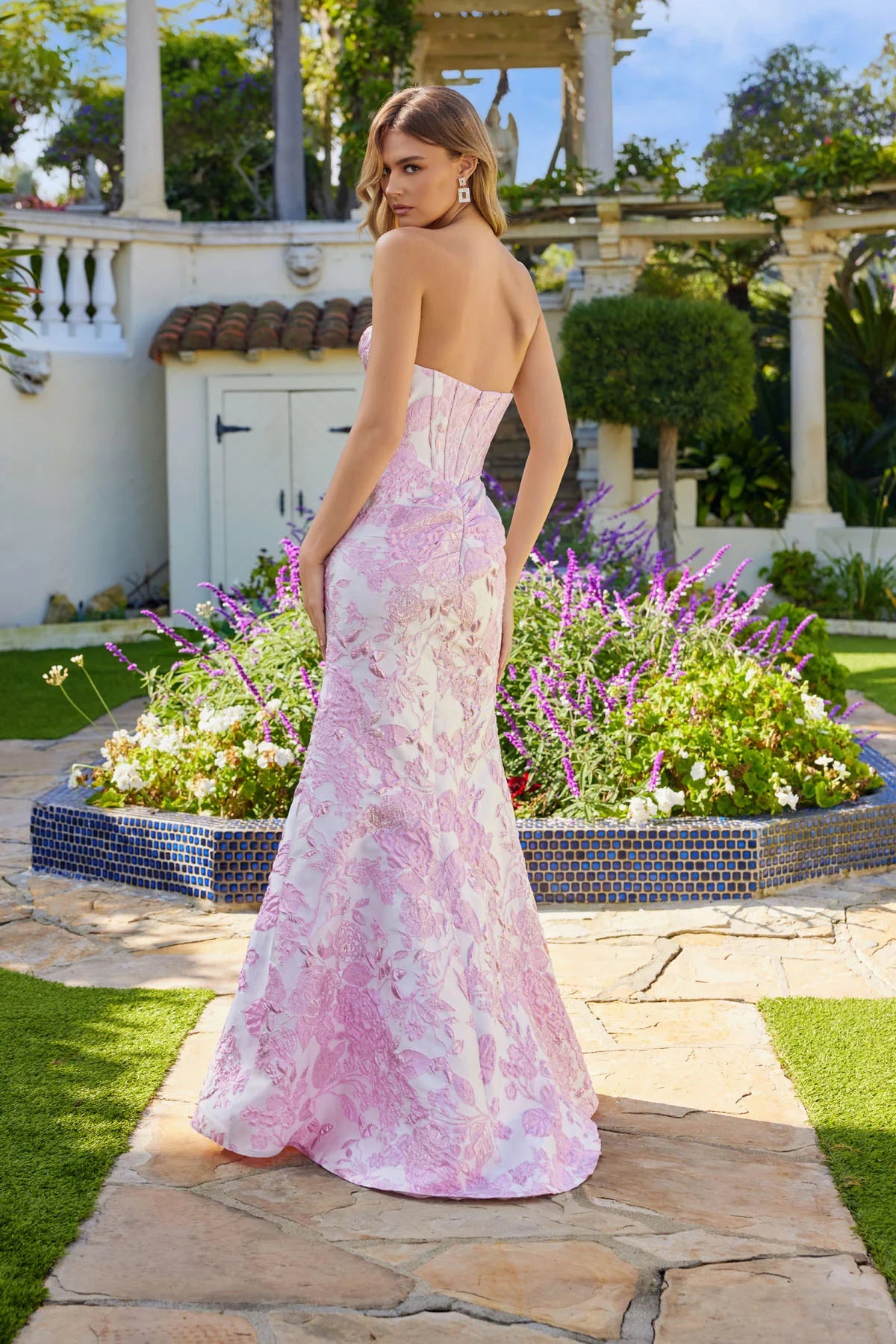 A woman stands outdoors in a strapless floral Long Prom Dress: Juliet JT2737A with a corset bodice, posing with her back to the camera as the pink and white floor-length gown flows elegantly before a lush garden backdrop.