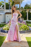 A woman stands outdoors in the Juliet JT2737A long prom dress in ivory pink, featuring a strapless floral design, corset bodice, and thigh-high slit, surrounded by flowers and elegant stone landscaping.