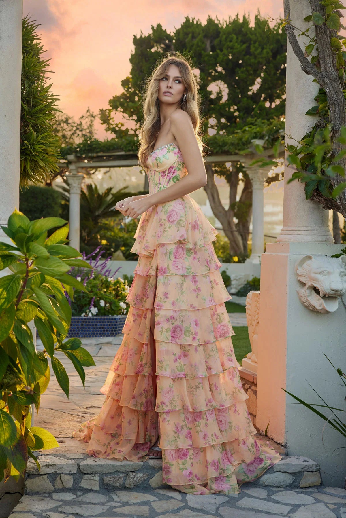 A woman stands outdoors at sunset in the Juliet JT2739S long prom dress, featuring a strapless floral design with a corset bodice and tiered chiffon A-line skirt, surrounded by greenery and white stone structures.