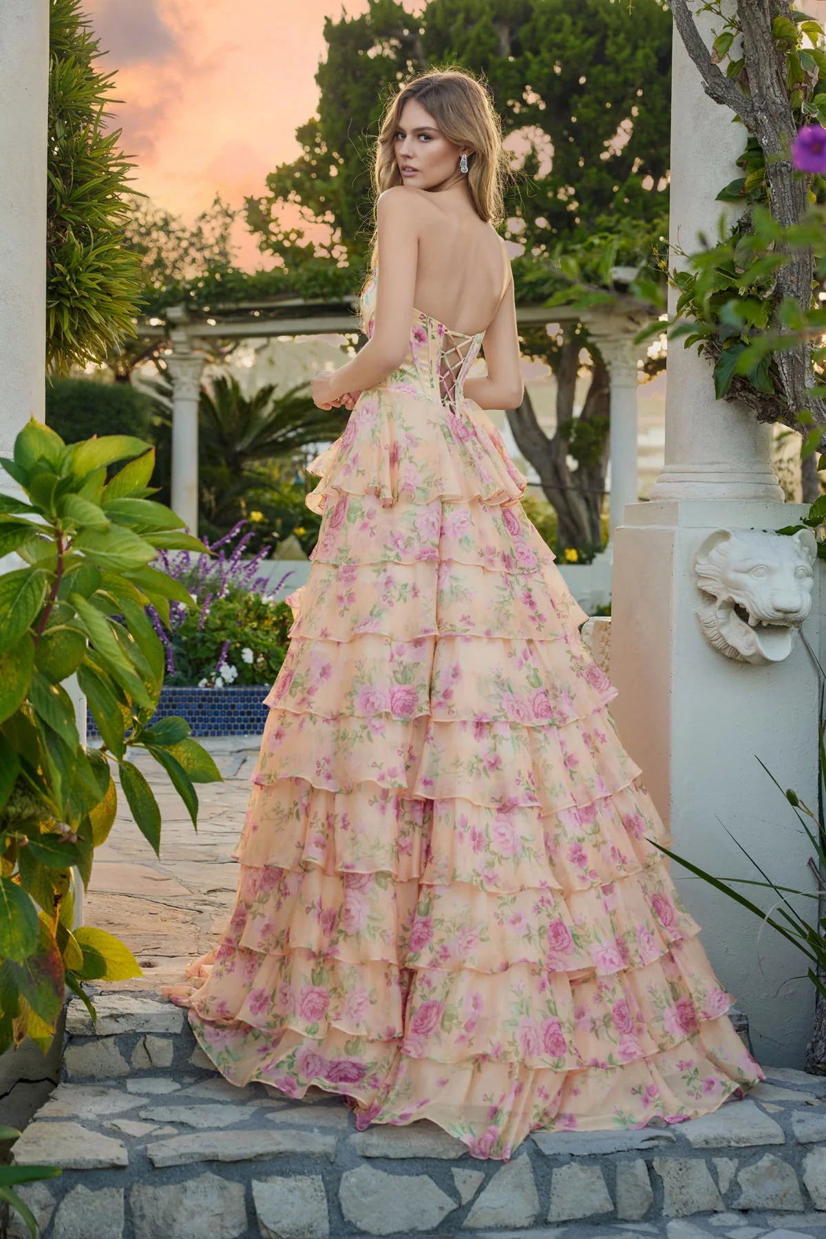 A woman stands outdoors on stone steps, wearing the Long Prom Dress: Juliet JT2739S, a strapless floral gown with a tiered chiffon A-line skirt in pink and green. Lush greenery and a sunset sky complete the scenic background.