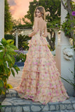 A woman stands outdoors on stone steps, wearing the Long Prom Dress: Juliet JT2739S, a strapless floral gown with a tiered chiffon A-line skirt in pink and green. Lush greenery and a sunset sky complete the scenic background.