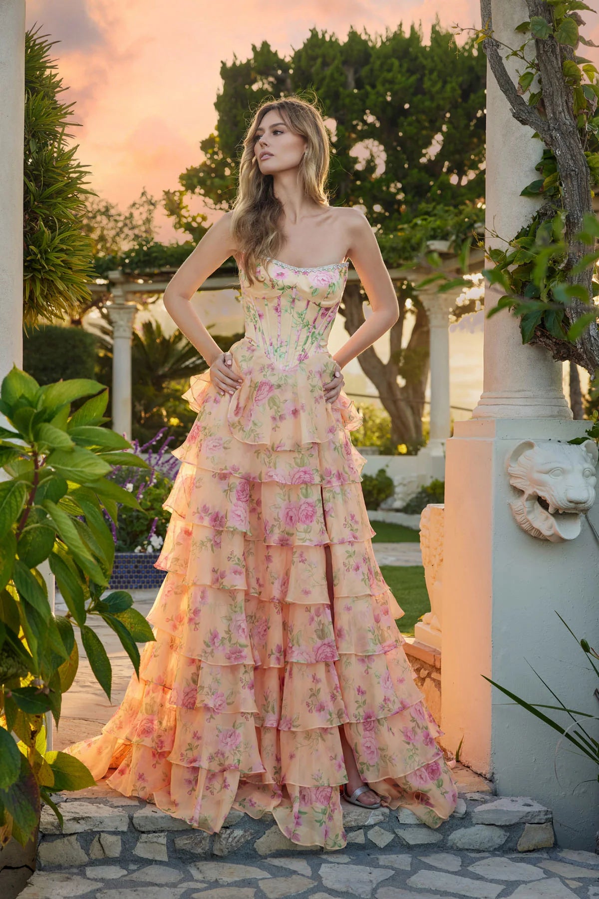 A woman stands outdoors at sunset before a garden pergola, wearing the Juliet JT2739S Long Prom Dress in Pastel Yellow Floral, featuring a strapless corset bodice and a flowing, ruffled chiffon A-line skirt with pink flowers.