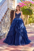 A woman stands outdoors in sunlight wearing the Long Prom Dress: Juliet JT2611N, a dark blue strapless gown with a fitted bodice, surrounded by stone walls and blooming flowers.