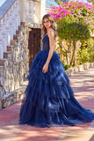 A woman stands outdoors in the Juliet JT2611N Long Prom Dress in navy blue, featuring a tiered tulle skirt and a beaded corset bodice, near a stone staircase and blooming flowers.