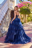 A woman stands outdoors on a stone path near flowers, wearing the Long Prom Dress: Juliet JT2611N, a dark blue tiered glitter tulle ball gown with a beaded corset bodice.