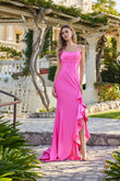 A woman stands outdoors on a stone path in the Juliet JT2727H Long Prom Dress in fuchsia, featuring ruffle detailing and a lace-up back, with greenery and a white building behind her.