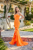 A woman stands outdoors in the Juliet JT2727H long prom dress in orange, featuring a form-fitting jersey design, ruffle details, spaghetti straps, and a lace-up back, surrounded by lush greenery and elegant stone architecture.
