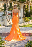  woman wears the Juliet JT2727H long prom dress in orange, standing outdoors on a stone path with a white pergola and greenery in the background, looking over her shoulder.