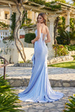 A woman wearing the Juliet JT2727H Long Prom Dress—a fitted, light blue jersey gown with a lace-up back and floor-length train—stands outdoors on a stone pathway, glancing over her shoulder.