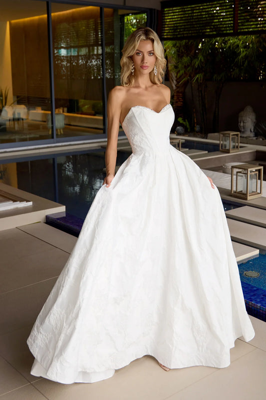 Woman in a white strapless gown standing in an outdoor setting with a pool and modern building.