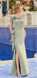 A woman stands by a pool in an olive Juliet JT2716C long prom dress, featuring elegant 3D rosettes, with flowers and a Mediterranean-style building in the background.