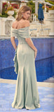 A woman stands by a pool in an olive Juliet JT2716C long prom dress, featuring elegant 3D rosettes, with flowers and a Mediterranean-style building in the background.