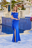 A woman in the Juliet JT2716C long prom dress in royal blue, featuring an off-the-shoulder design, side slit, and 3D rosette detail, stands by a blue-tiled pool in a stylish outdoor setting.