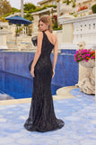 A woman wearing the Juliet JT2723C long prom dress with a black one-shoulder neckline stands by a blue-tiled pool outdoors, surrounded by floral arrangements and classical architecture, facing away.