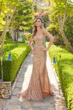 A woman stands on a stone garden path, surrounded by greenery and sunlight, wearing the Juliet JT2730H Long Prom Dress with thin straps, sparkling sequins, and a high leg slit.