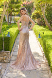 A woman stands on a stone garden path wearing the Juliet JT2730H, a sparkling floor-length sequin prom dress with thin straps and a backless design, surrounded by trees, bushes, and lamps.