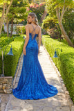 Wearing the Juliet JT2730H long prom dress—a sparkly blue sequin gown with a backless, crisscross strap design—a woman stands on a stone garden path bordered by trimmed hedges and outdoor lamps.