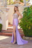 A woman wears the Juliet JT2729H long prom dress, featuring a strapless beaded bodice and thigh-high slit, while standing outdoors on tiled steps near green plants and stone owl statues.