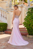 A woman in the Juliet JT2729H long prom dress, featuring a strapless beaded bodice and leg slit, stands outdoors on a tiled patio surrounded by greenery and ornate stone structures, facing away.