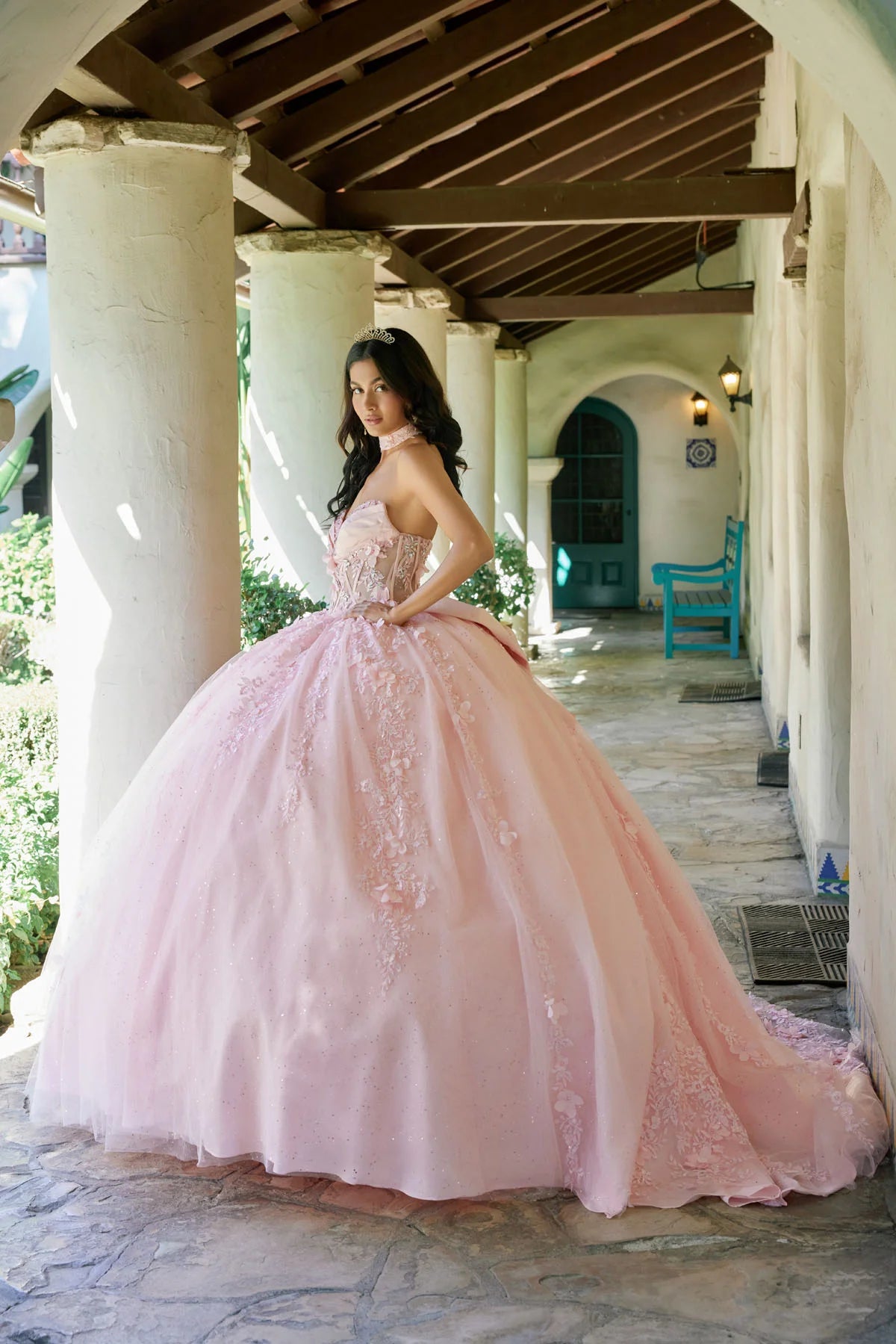 Woman in a pink ball gown standing in a sunlit outdoor setting with columns and plants.