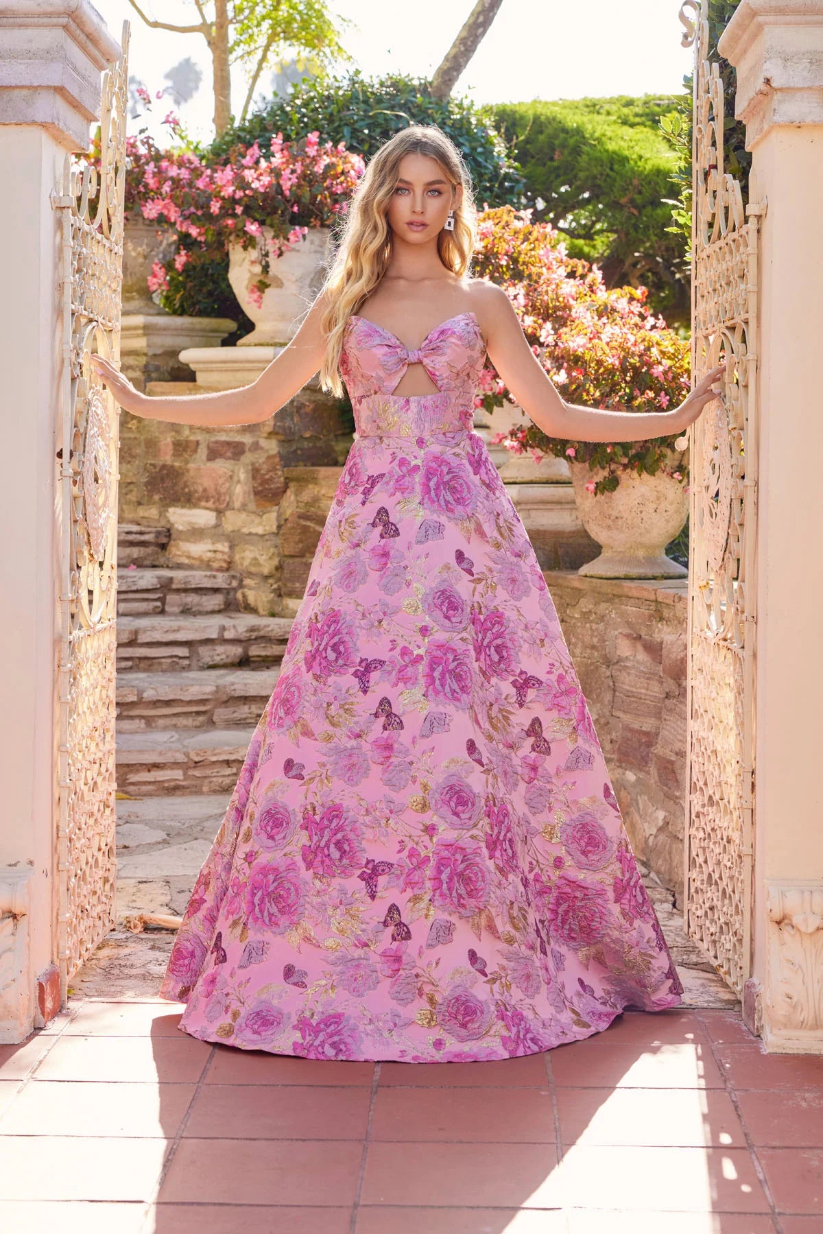 Wearing the Juliet JT2735H long prom dress, a woman stands by an open wrought iron gate, stone steps and blooming flowers behind her.