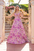 Wearing the Juliet JT2735H long prom dress, a woman stands by an open wrought iron gate, stone steps and blooming flowers behind her.