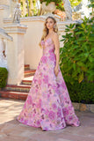 A woman in a Pink Floral Juliet JT2735H Long Prom Dress stands outside by steps, with greenery and decorative statues in the background.