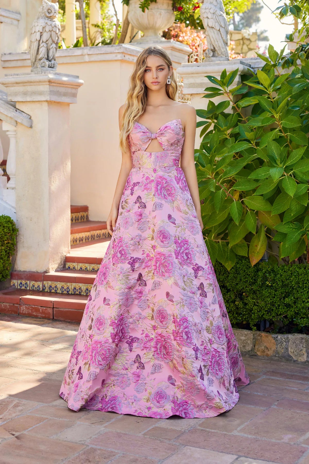 A woman stands outdoors in the Juliet JT2735H long prom dress, featuring a strapless floral design and cutout bodice, surrounded by greenery and architectural details.