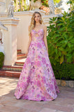 A woman stands outdoors in the Juliet JT2735H long prom dress, featuring a strapless floral design and cutout bodice, surrounded by greenery and architectural details.
