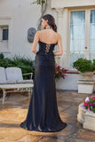 A woman wearing the Juliet JT2698A long black satin prom dress with a strapless corset bodice and side lace-up detail stands on a stone patio near a garden and white building.