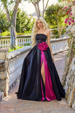 Wearing the Juliet JT2733H, a strapless black long prom dress with a pink rose accent and pink inner lining, a woman stands on a stone pathway surrounded by greenery.