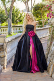 A woman with long blonde hair models the Juliet JT2733H Long Prom Dress—black, strapless, with a high slit, bright pink lining, and a large pink rose at the waist—while standing on a garden stone path.