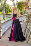 A woman poses outdoors on a stone walkway in the Juliet JT2733H long prom dress, featuring a strapless black silhouette with a fuchsia front panel, bow, and subtle 3D floral accents.