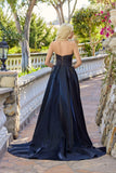 A blonde woman stands outdoors on a stone path, wearing the Juliet JT2733H—a strapless black organza a-line prom dress with a lace-up back and sweeping train.
