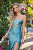 Wearing the Juliet JT2708K long prom dress, a woman in teal satin with an embellished strapless corset bodice stands outdoors beside a lush garden filled with greenery and pink flowers.