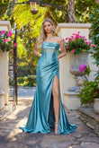 A woman stands outdoors by a stone planter with pink flowers and greenery, wearing the Juliet JT2708K long prom dress—a teal satin strapless gown with a wrap-style sheath skirt and thigh-high slit.