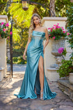 A woman stands outdoors by a stone planter with pink flowers and greenery, wearing the Juliet JT2708K long prom dress—a teal satin strapless gown with a wrap-style sheath skirt and thigh-high slit.
