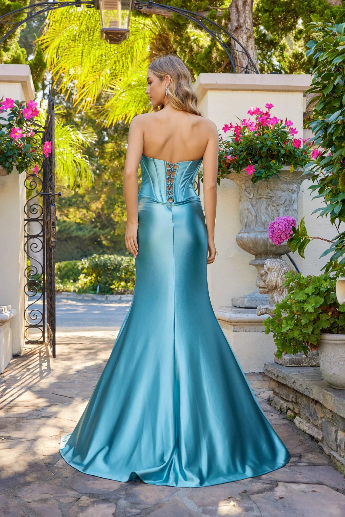 Wearing the Long Prom Dress: Juliet JT2708K, a woman stands outdoors on a stone path amid greenery and pink potted flowers. The satin strapless dress features an embellished corset bodice.