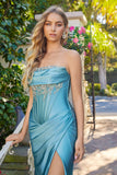 Wearing the Juliet JT2708K long prom dress, a woman in teal satin with an embellished strapless corset bodice stands outdoors beside a lush garden filled with greenery and pink flowers.
