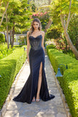 Wearing the Long Prom Dress Juliet JT2706K, a woman poses in a manicured garden. The strapless black gown features an embellished corset bodice and a high slit as she stands on a stone path.